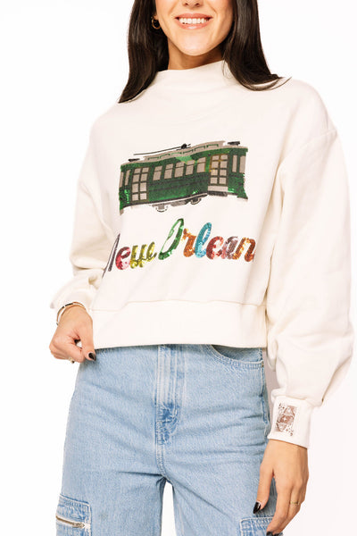 “Queen of Sparkles light beige sweatshirt with sequin yellow taxi and rainbow script lettering, mock-neck design, and relaxed fit.”